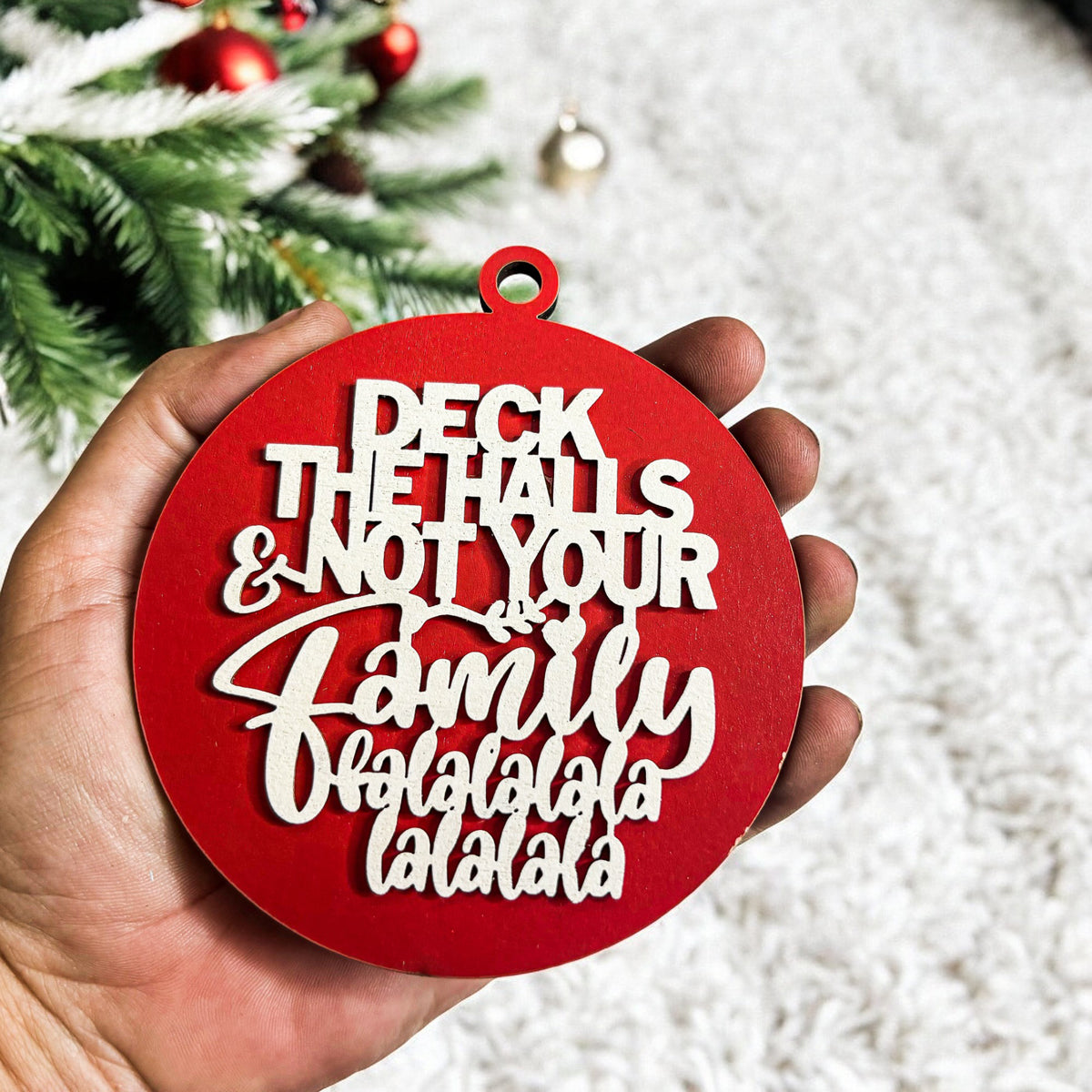 Red Christmas ornament with humorous text held in front of a decorated tree.